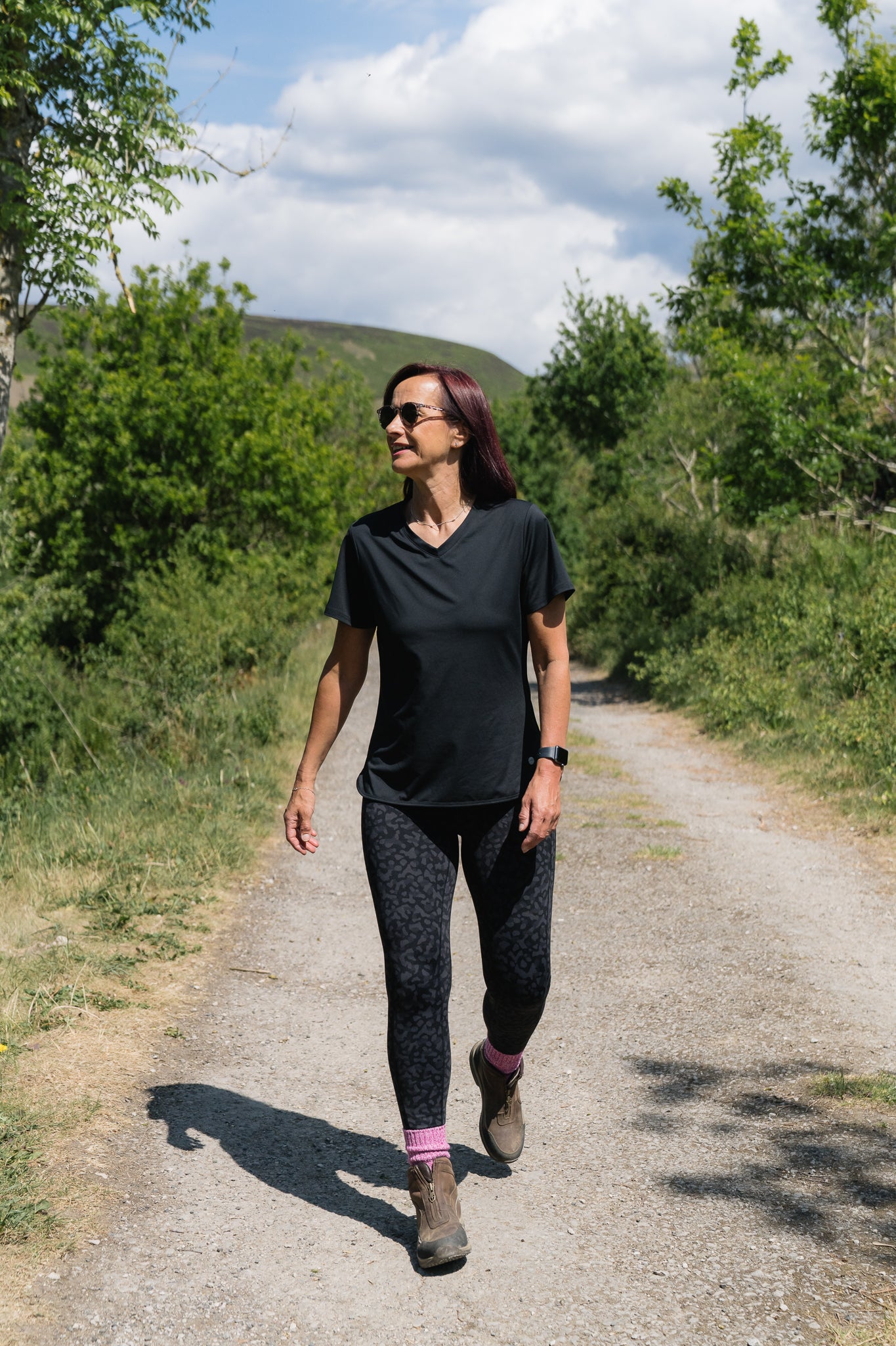 Woman walking on a dirt path in a natural setting with trees and mountains in the background.