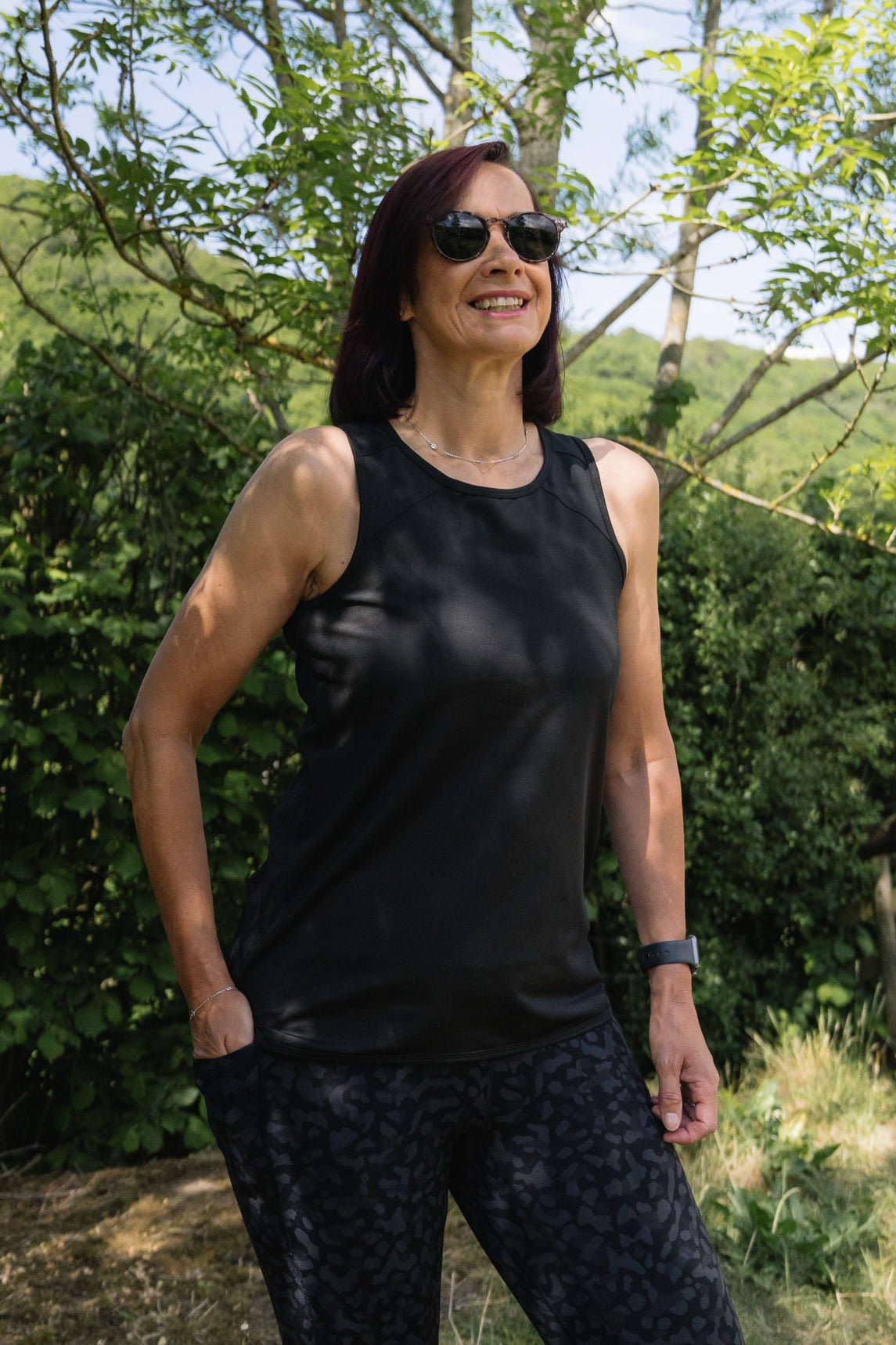 Woman wearing a black tank top and sunglasses standing outdoors with greenery in the background