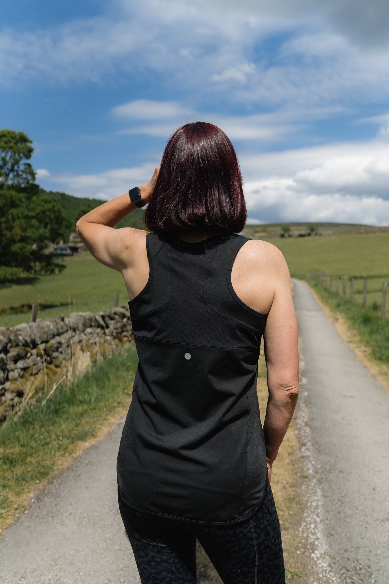 Person in a black sleeveless sports top standing on a country road with green fields and trees in the background.