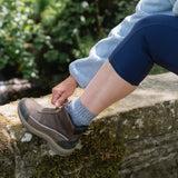 Person wearing brown shoes and blue socks sitting on a stone ledge with greenery in the background