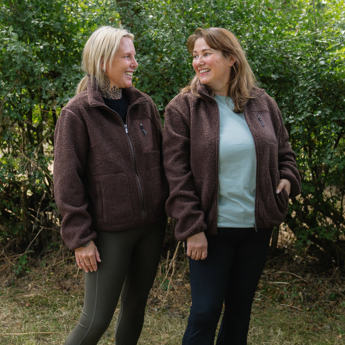 Two women standing outdoors in front of a green hedge, wearing matching brown sherpa zip-up jackets. One woman has blonde hair and wears dark olive leggings, while the other has light brown hair and wears dark blue leggings over a light blue top.