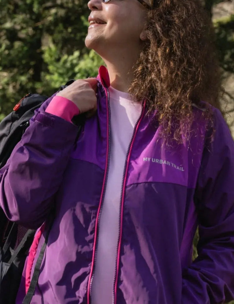 A person wearing a purple and pink waterproof jacket with a hood, holding a matching packable drawstring bag, standing outdoors.