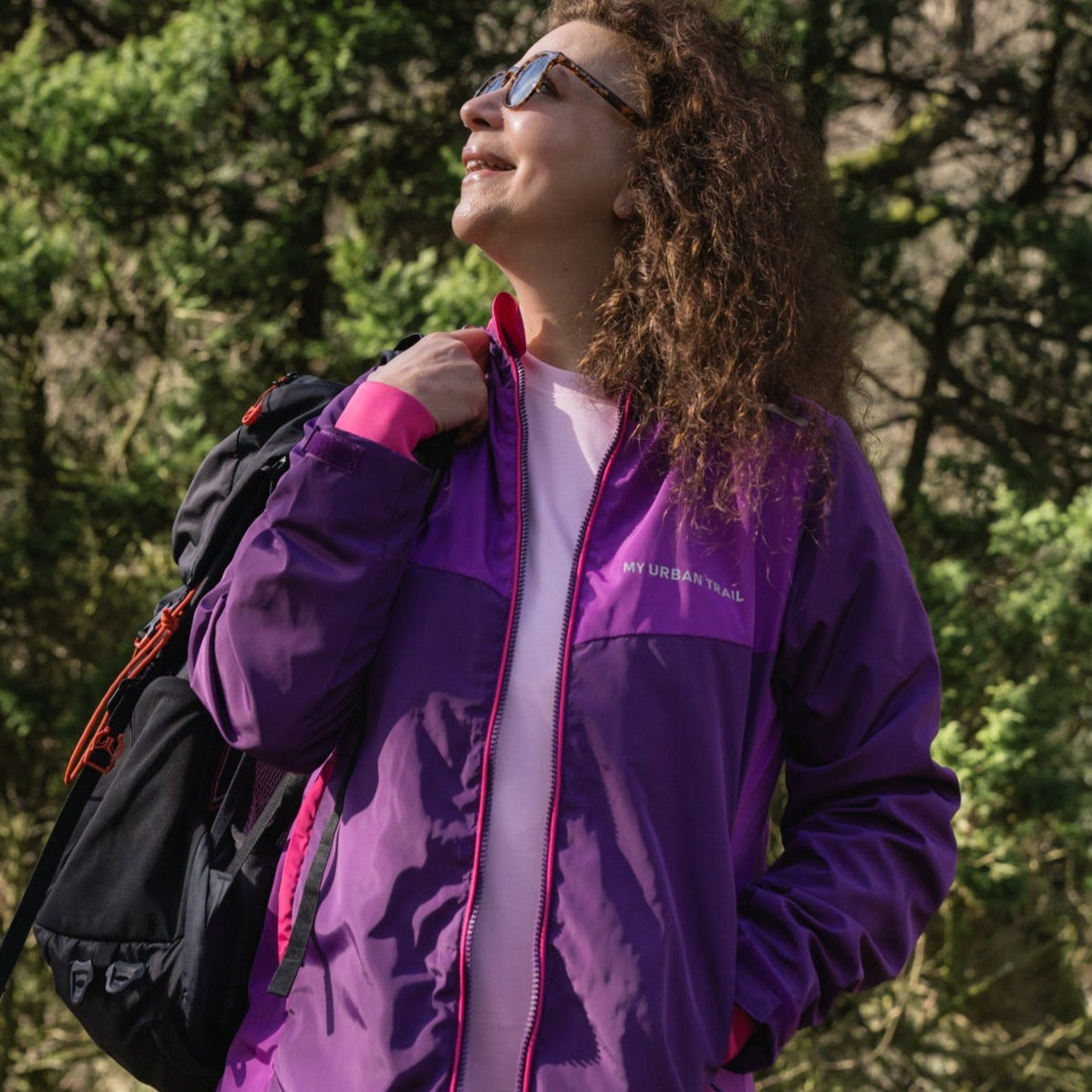 A person wearing a purple and pink waterproof jacket with a hood, holding a matching packable drawstring bag, standing outdoors.