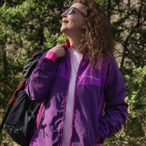 A person wearing a purple and pink waterproof jacket with a hood, holding a matching packable drawstring bag, standing outdoors.