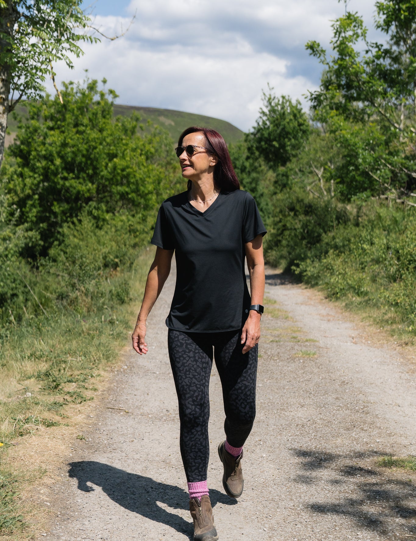 Woman walking on a dirt path in a natural setting with trees and mountains in the background.