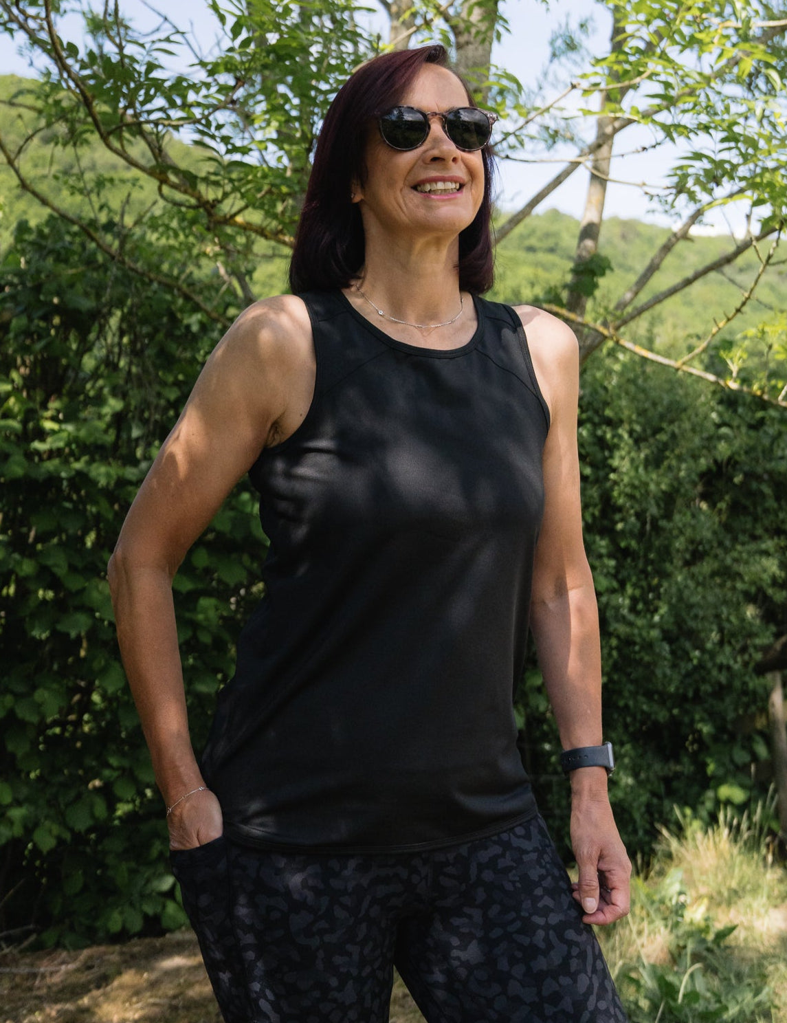 Woman wearing a black tank top and sunglasses standing outdoors with greenery in the background