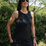 Woman wearing a black tank top and sunglasses standing outdoors with greenery in the background