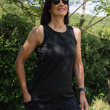 Woman wearing a black tank top and sunglasses standing outdoors with greenery in the background