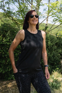 Woman wearing a black tank top and sunglasses standing outdoors with greenery in the background