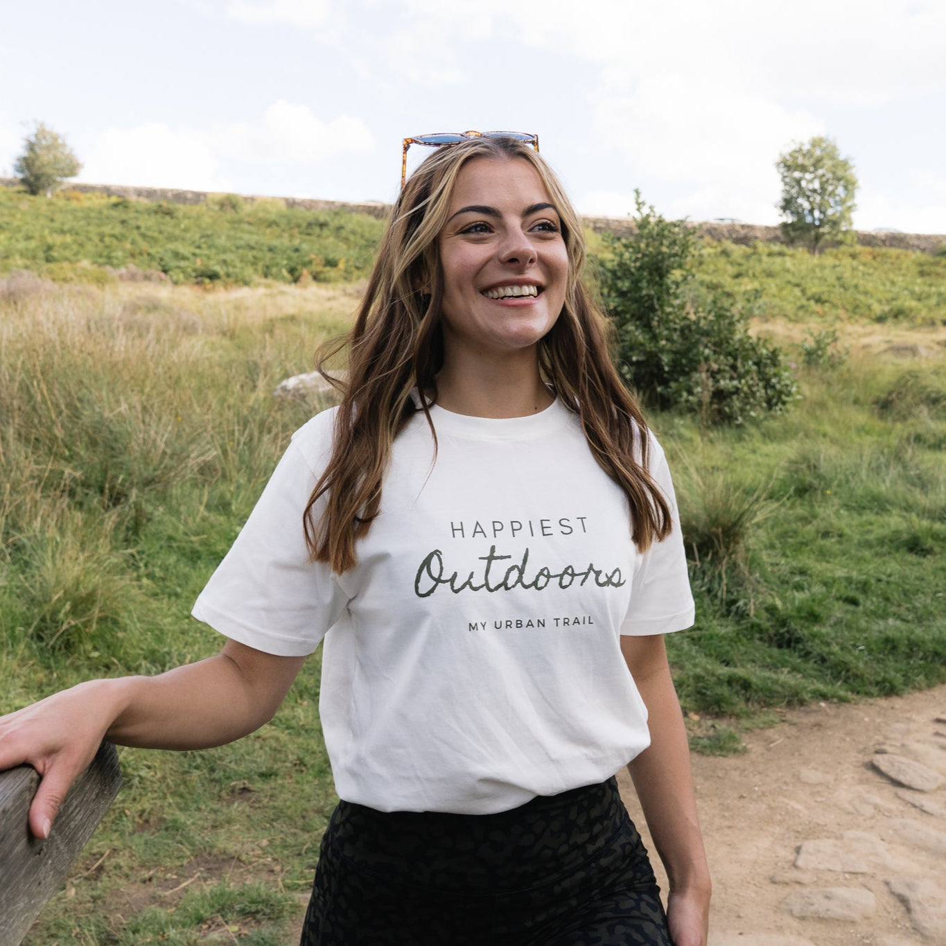 Woman wearing a 'Happiest Outdoors' t-shirt standing in a grassy field.