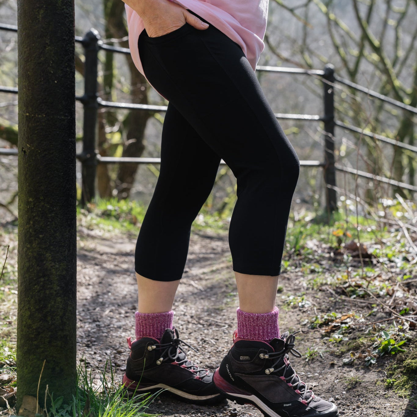 A person wearing black capri hiking leggings with a pink top and walking shoes standing next to a fence in a natural outdoor setting.
