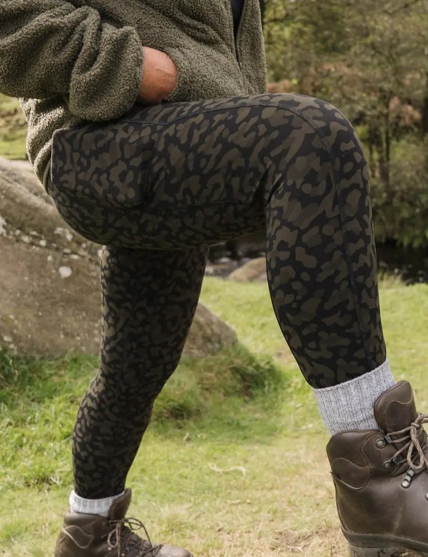 A person wearing high-waisted khaki print hiking leggings with a thick waistband, standing outdoors.