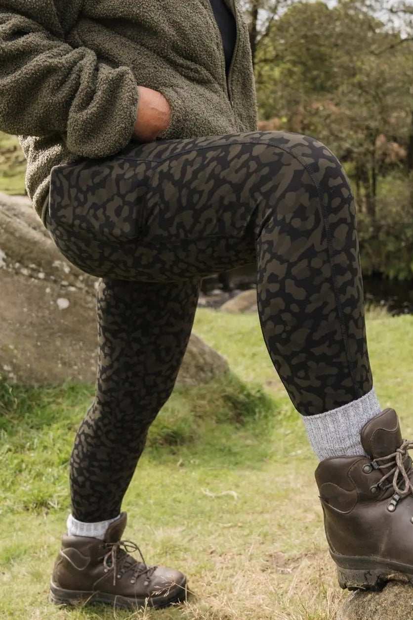A person wearing high-waisted khaki print hiking leggings with a thick waistband, standing outdoors.