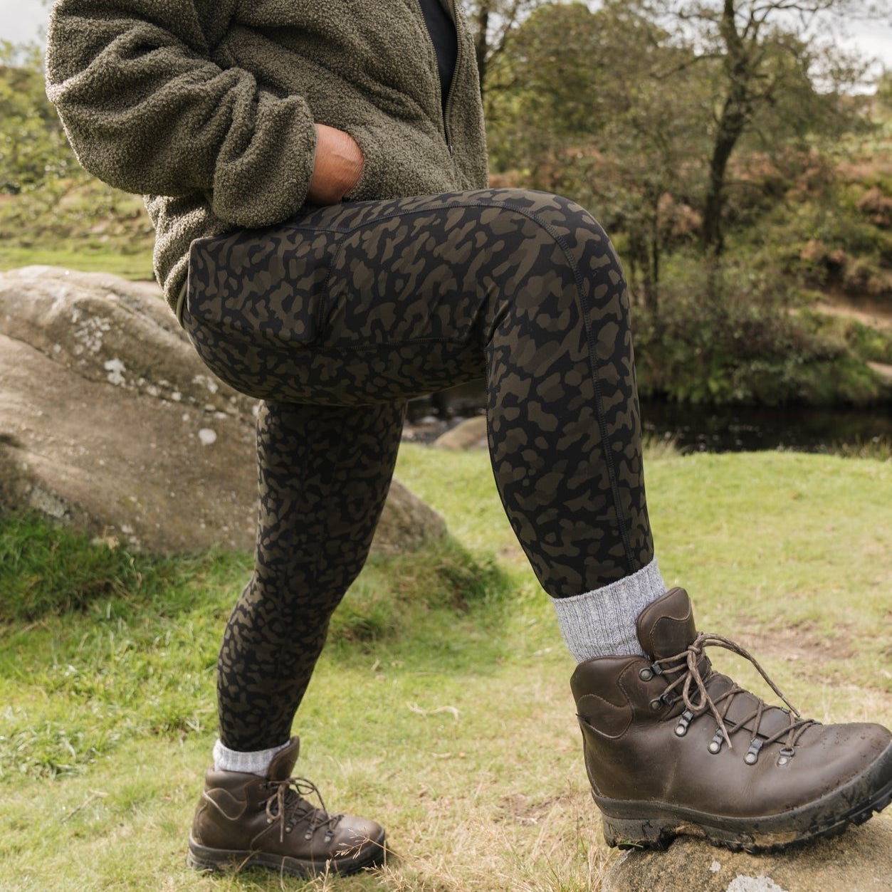 A person wearing high-waisted khaki print hiking leggings with a thick waistband, standing outdoors.