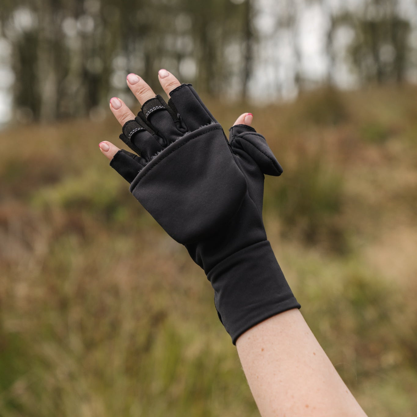 A person wearing black fingerless hiking mittens with magnetic fastenings, in an outdoor setting.