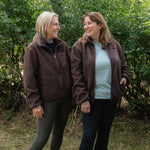 Two women standing outdoors in front of a green hedge, wearing matching brown sherpa zip-up jackets. One woman has blonde hair and wears dark olive leggings, while the other has light brown hair and wears dark blue leggings over a light blue top.