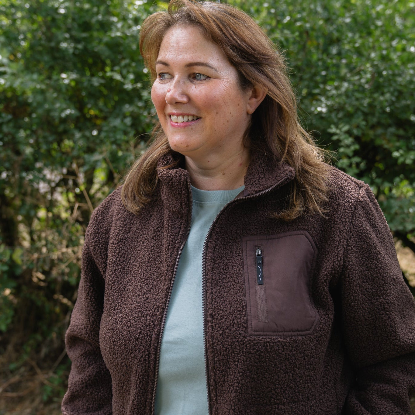 A woman with light brown hair smiling outdoors, wearing a brown fleece zip-up jacket over a light blue top, with green foliage in the background.