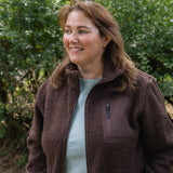 A woman with light brown hair smiling outdoors, wearing a brown fleece zip-up jacket over a light blue top, with green foliage in the background.