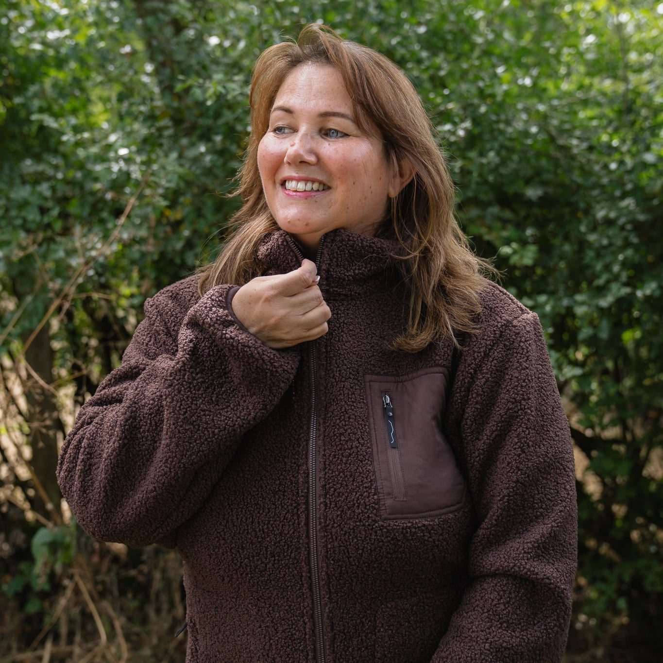 A woman wearing a brown fleece zip-up jacket stands outdoors in front of a green hedge, smiling and adjusting the collar.