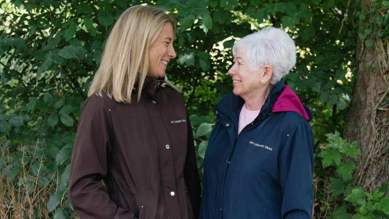 Two women standing outdoors in front of a lush green tree, wearing waterproof jackets. The woman on the left wears a dark brown hooded jacket, and the woman on the right wears a navy blue hooded jacket with a pink inner hood lining.